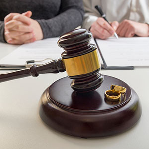 A gavel and two wedding rings rest on a desk where two people are signing documents.
